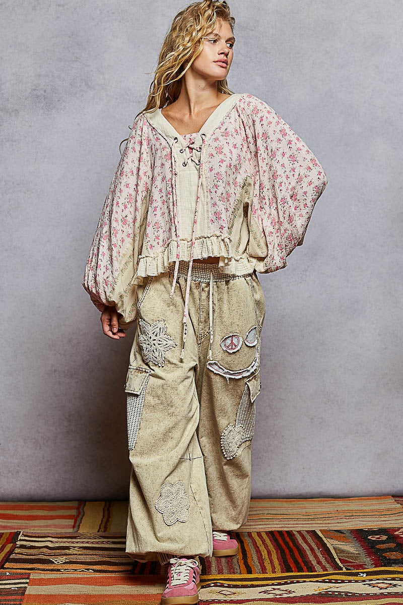 Woman wearing a floral top and beige pants with embroidery, standing on a colorful rug against a gray wall.