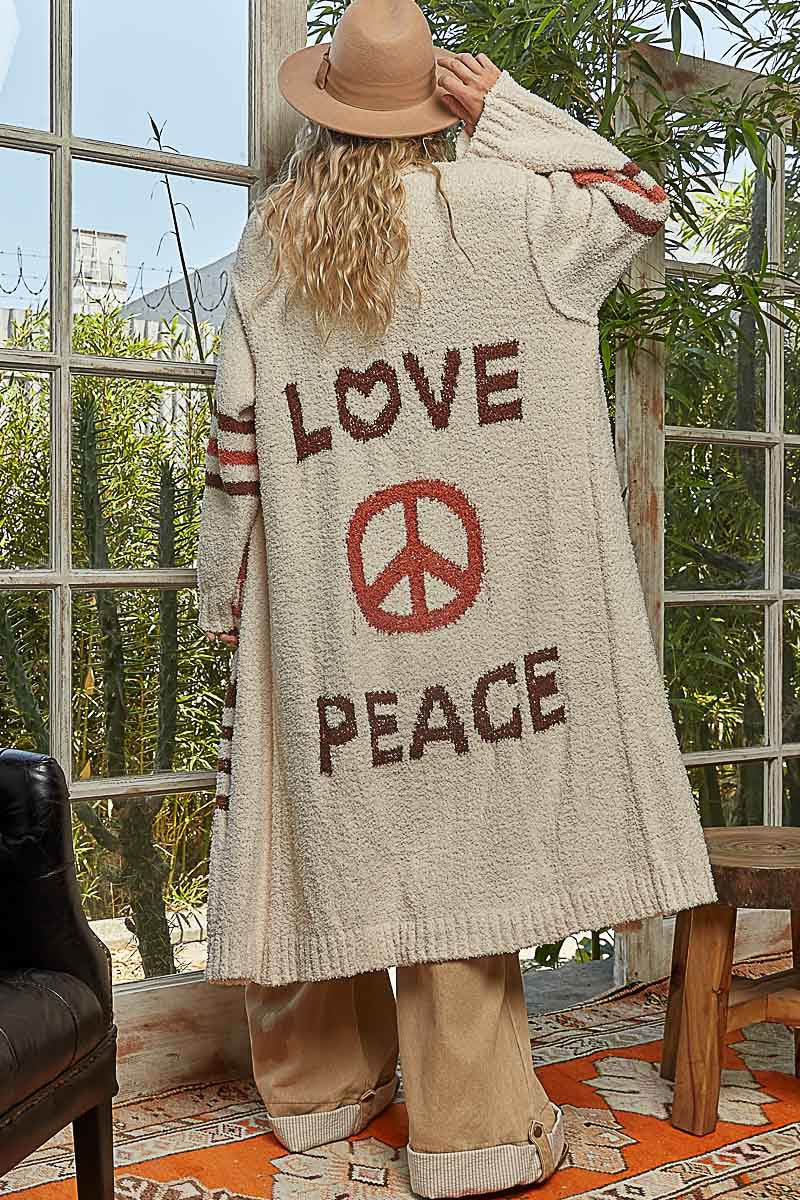 Person wearing a long beige cardigan with 'LOVE' and 'PEACE' text and symbols, standing in a room with plants and a window.