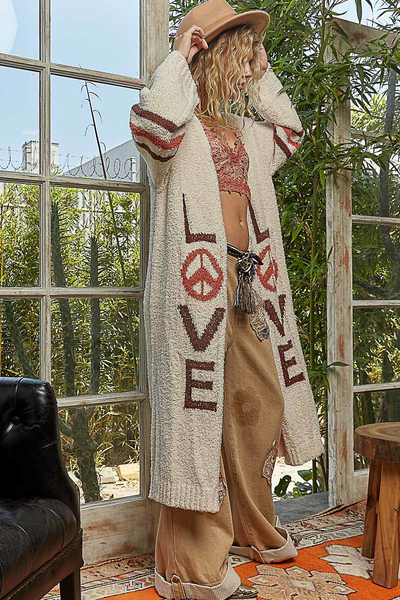 Person wearing a long, patterned cardigan with 'LOVE' and peace symbol in an outdoor setting.