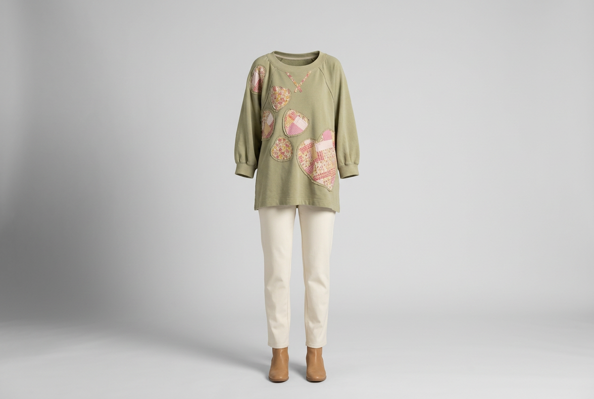 Sage sweatshirt with pink patchwork hearts, cream trousers and tan ankle boots against a shaded gray studio backdrop.