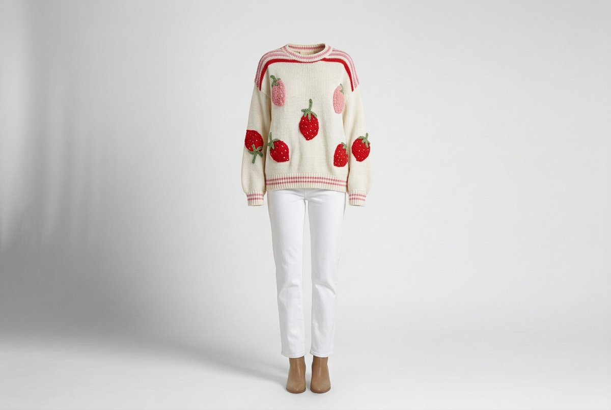 Ivory strawberry-appliqué sweater with pink striped trim, white jeans and tan ankle boots against a shaded gray backdrop.