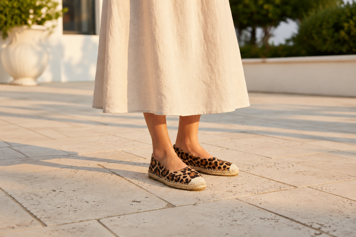 A person wearing a long cream linen skirt and leopard-print espadrille flats, standing on a sunlit stone patio.