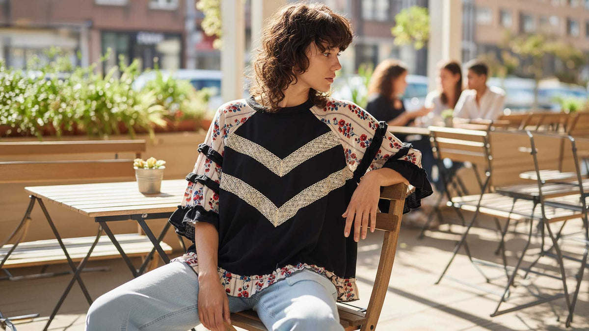 A woman sits on a wooden chair at an outdoor cafe table, wearing a black and cream chevron-patterned top with floral lace sleeves and light blue jeans.