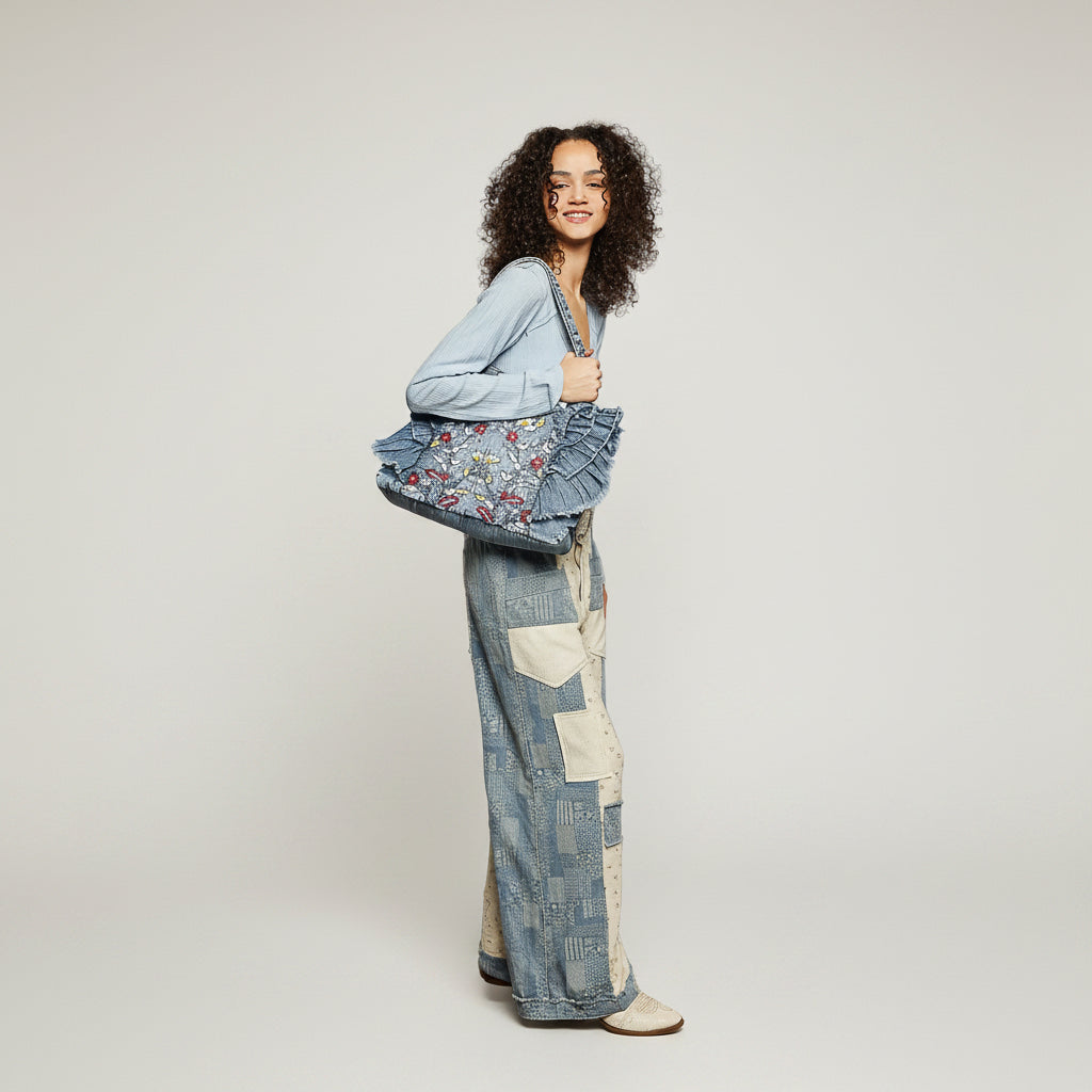 Women's blue denim floral embroidered tote bag with ruffle fringe trim worn on shoulder, close-up carry view