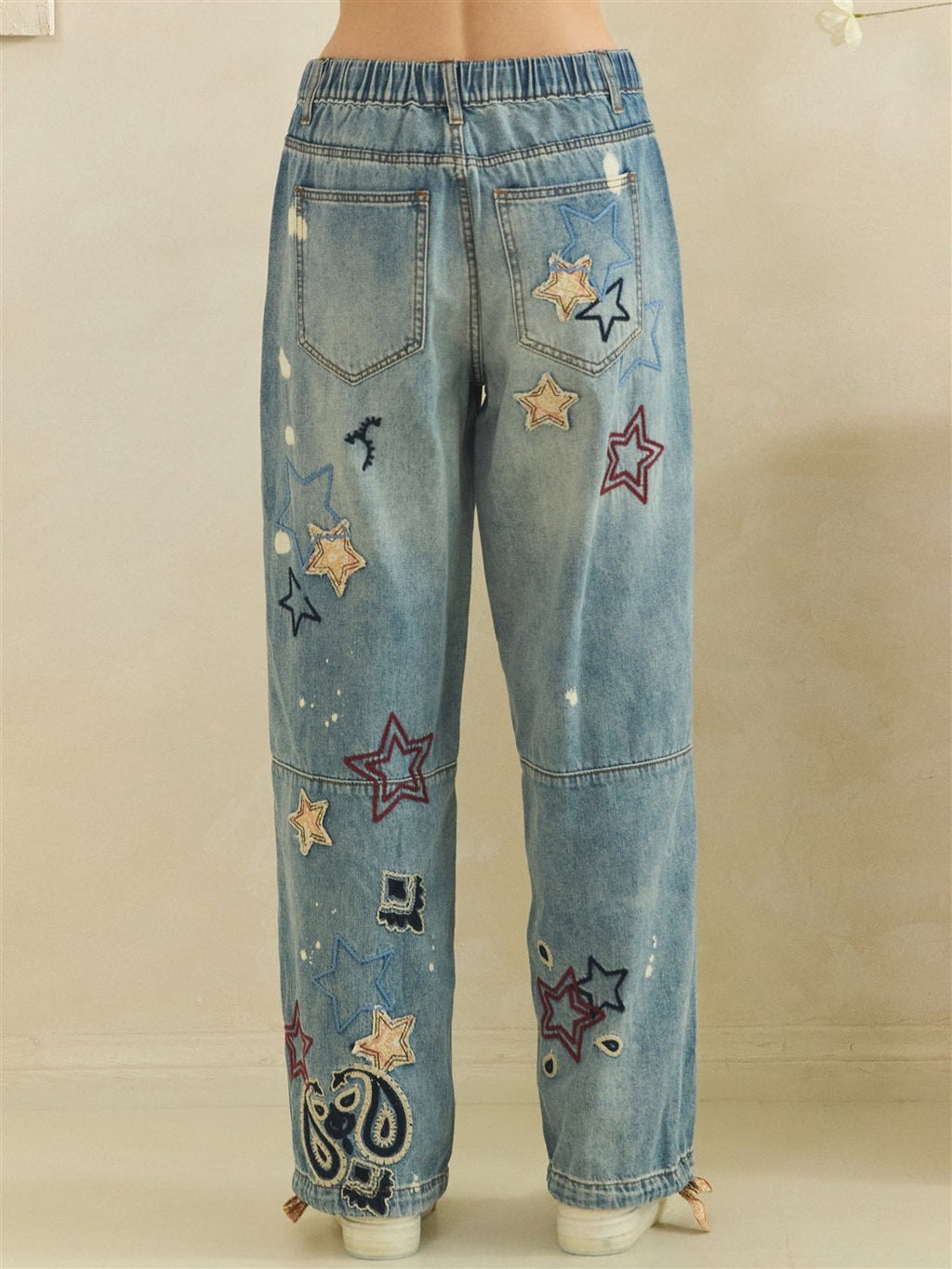 Back waist-to-hem view of blue jeans with patch pockets, scattered embroidered stars and paisley near the hem