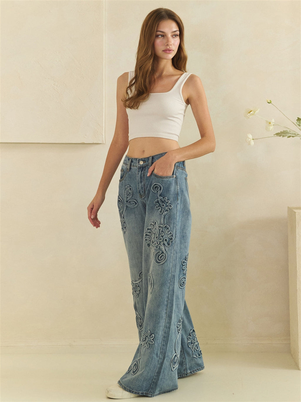 Three-quarter view of blue wide-leg jeans showing tonal flower appliques and curved front pockets.