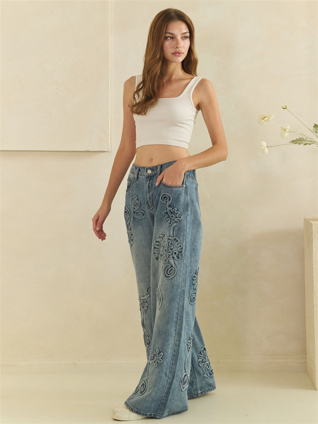 Three-quarter view of blue wide-leg jeans showing tonal flower appliques and curved front pockets.