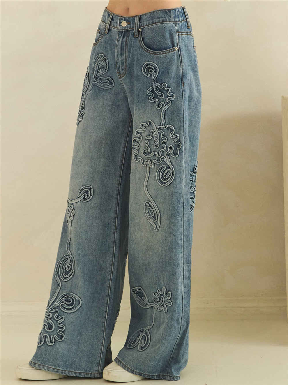 Angled waist-to-hem detail of washed blue jeans with floral appliques and broad leg openings.