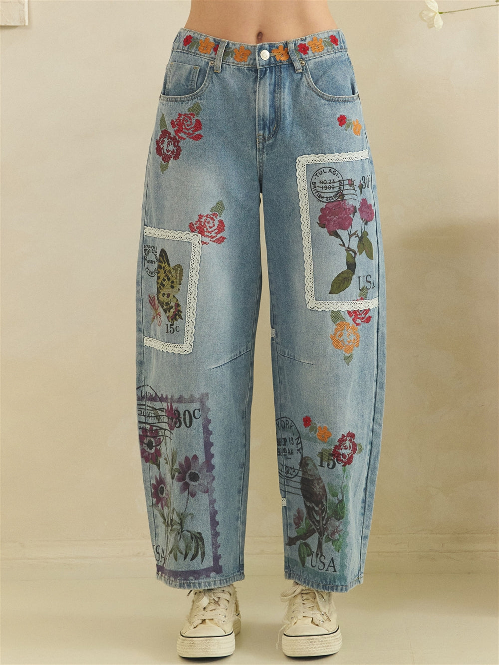 Front close view of blue barrel jeans with floral embroidery and lace-edged postage print panels
