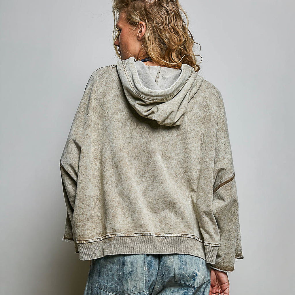 A woman viewed from behind wearing a faded light gray hooded sweatshirt and blue jeans against a plain light gray studio background.
