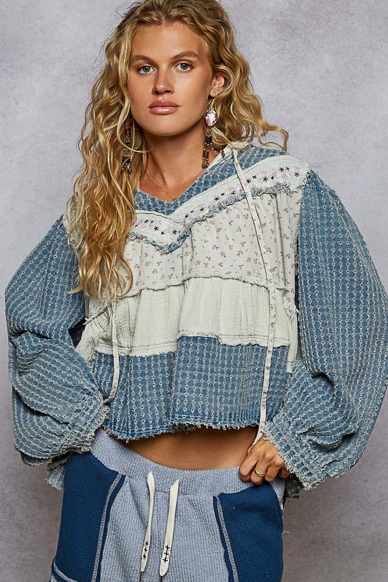 Close front view of a blue hooded pullover with embroidered trim, cream floral panels and long hood ties.