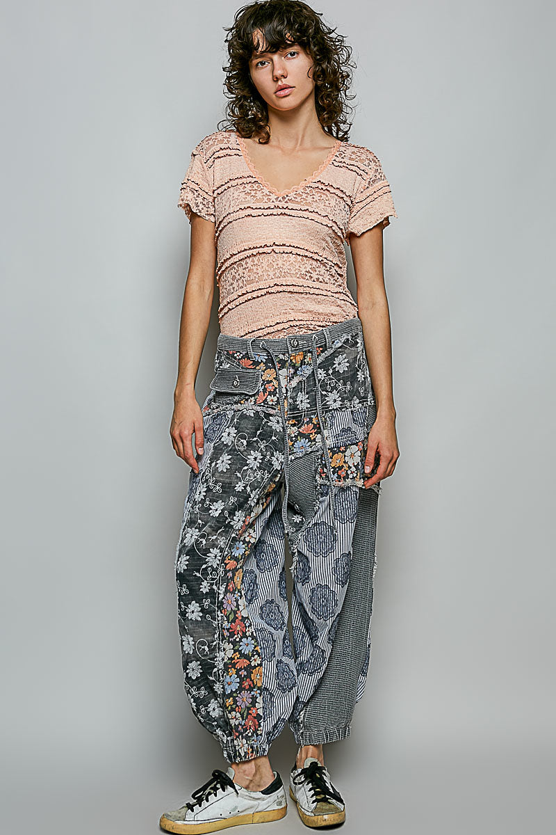 A model wearing a light pink textured short-sleeve top and gray patterned wide-leg pants with black and white sneakers, standing against a solid gray studio background.