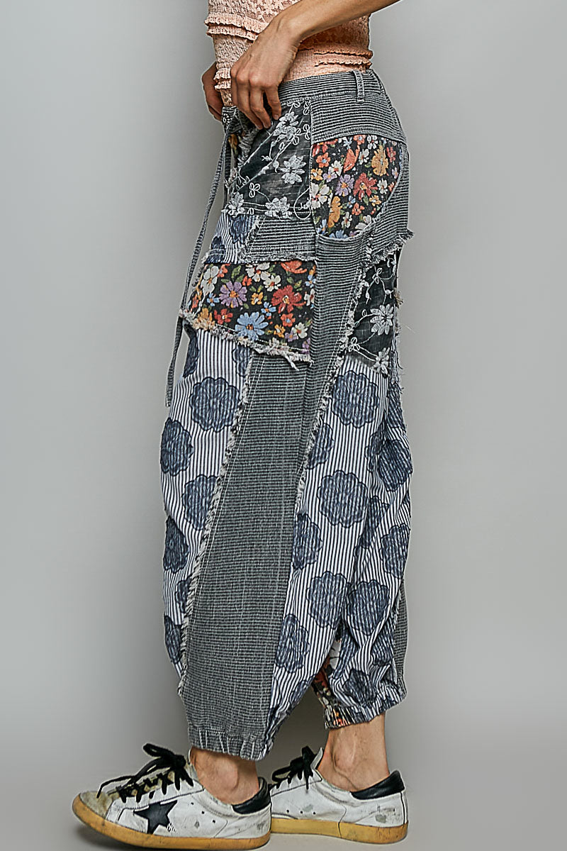 A person wearing loose, patchwork-style harem pants with mixed prints including floral, striped, and blue-gray patterned panels, paired with white sneakers against a plain light gray studio background.