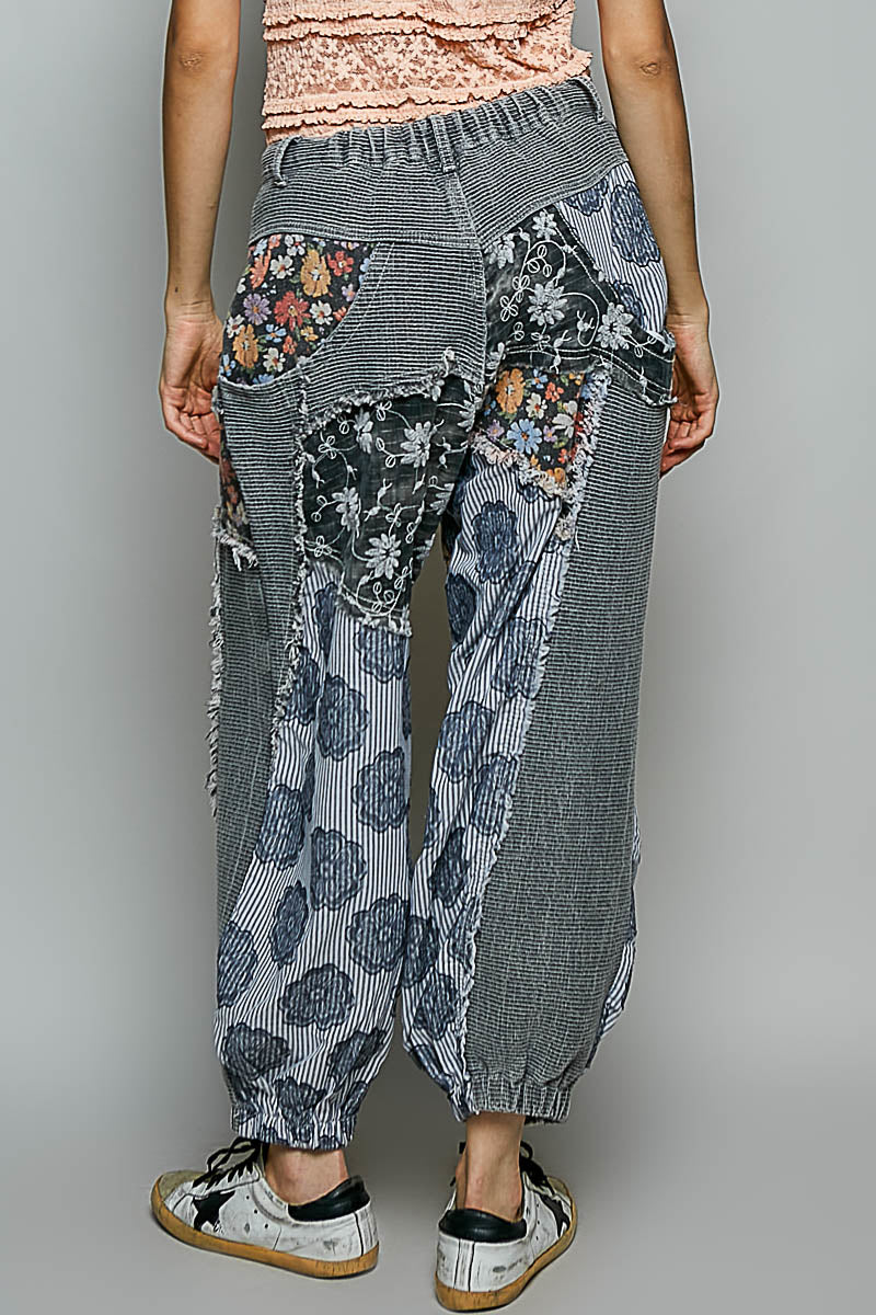 A person shown from the back wearing loose patchwork harem-style pants with gray striped and floral fabric panels, paired with white sneakers, against a plain light gray studio background.