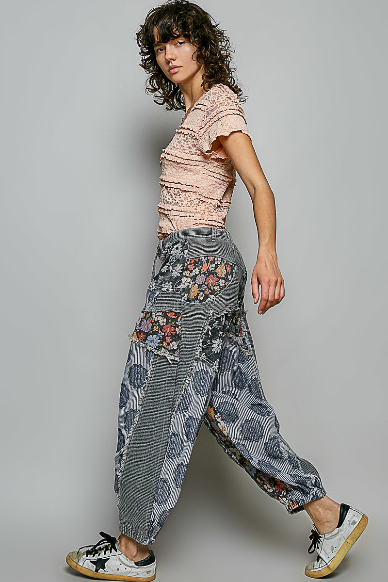 A model wearing a light pink textured short-sleeve top and high-waisted, wide-leg patchwork pants with floral and denim-like panels, paired with white sneakers, against a solid gray studio background.