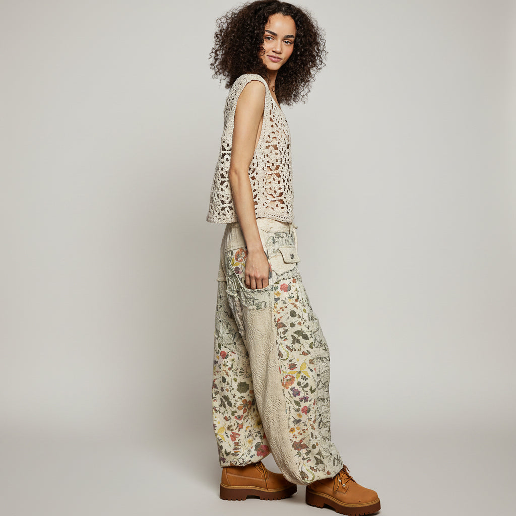 A woman modeling a sleeveless cream crochet vest and matching wide-leg floral pants with brown boots, standing against a textured light gray wall.