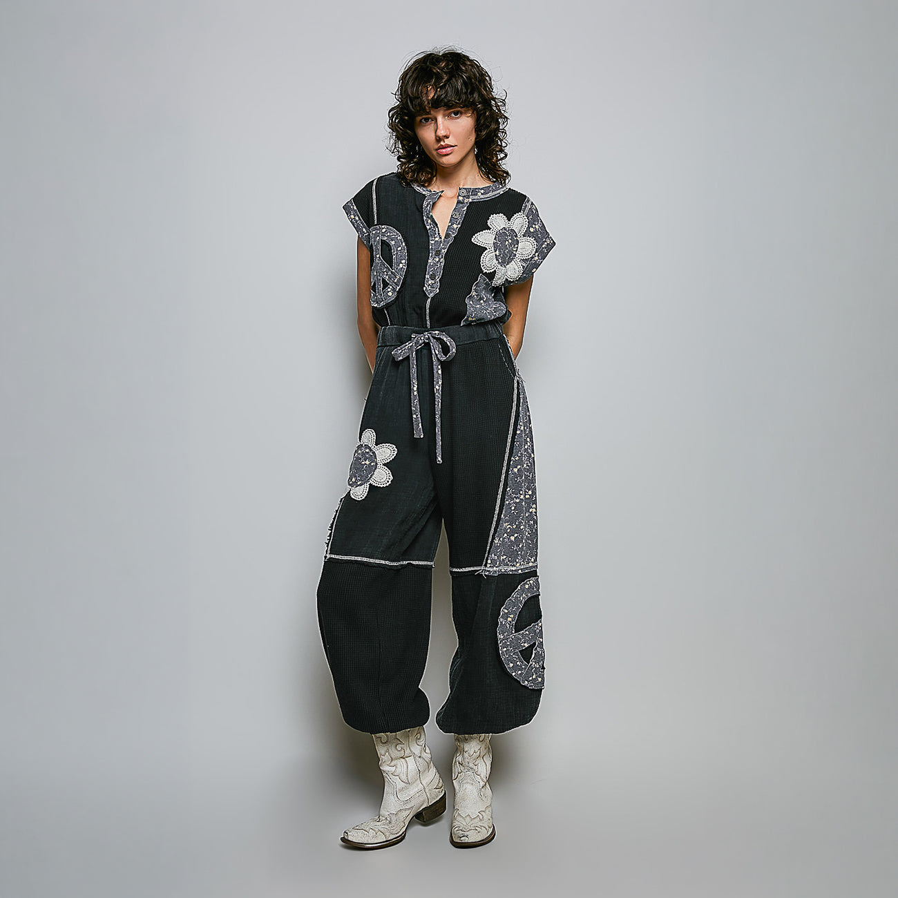 A model stands against a light gray studio background wearing a black wide-leg jumpsuit with silver paisley and floral prints, paired with white western-style boots.