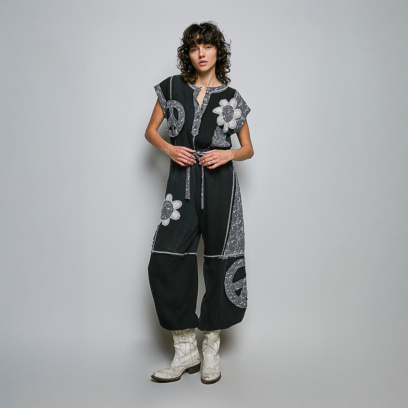 A model stands against a light gray studio background wearing a loose black jumpsuit with silver paisley and white floral applique, paired with white western boots.