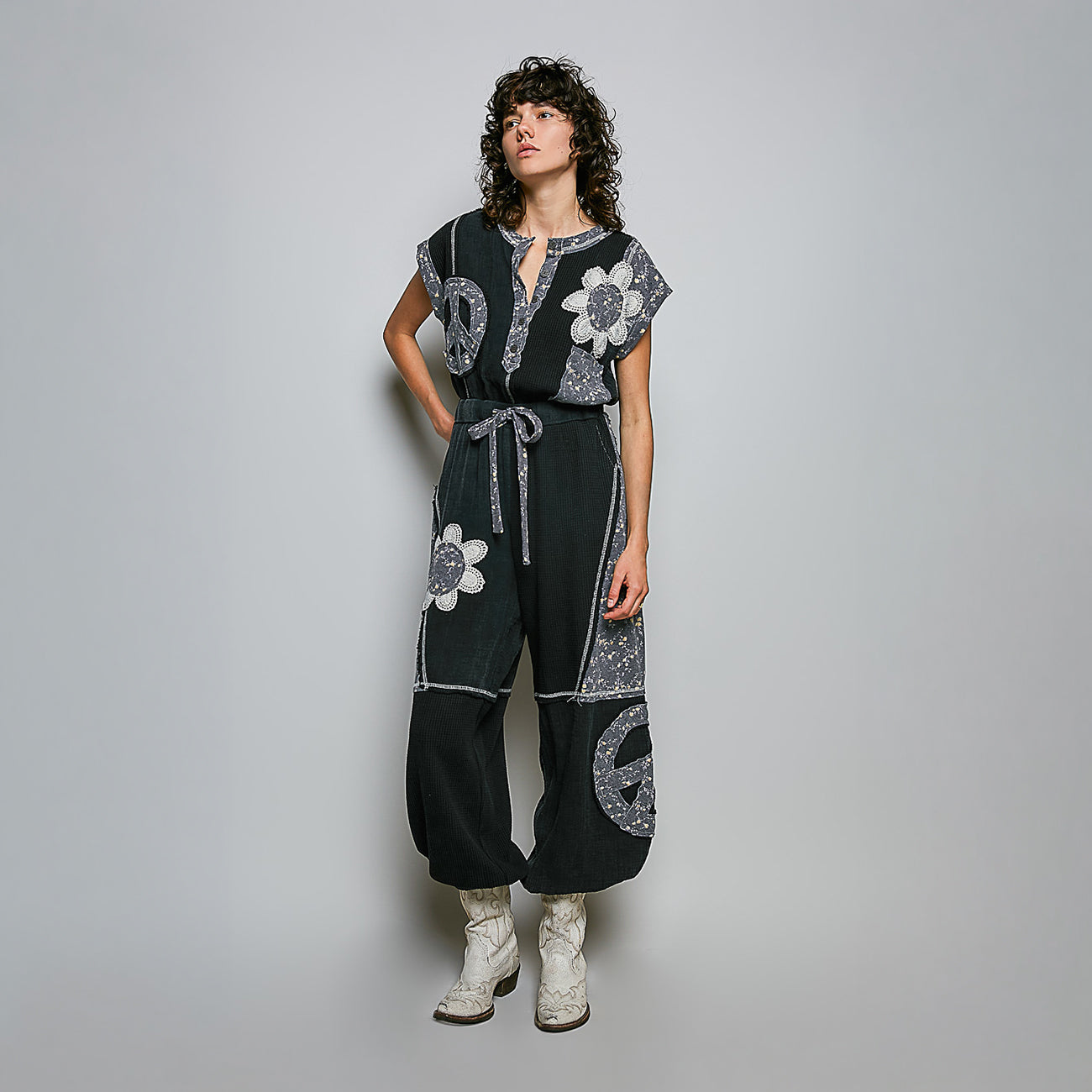 A model stands against a light gray studio background wearing a black wide-leg jumpsuit with silver-gray floral and circular patchwork details, paired with white lace-up boots.