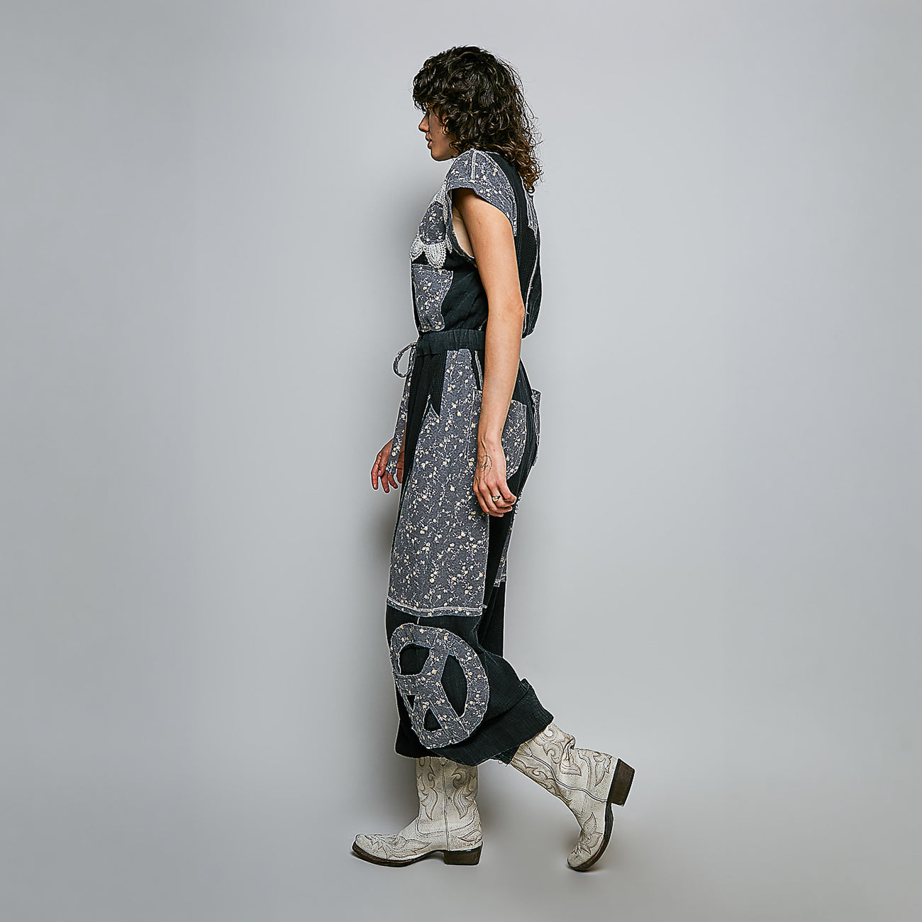 A model is shown from the side wearing a long, dark gray and black patterned dress with a peace sign motif on the leg, paired with white western boots, against a solid light gray studio background.