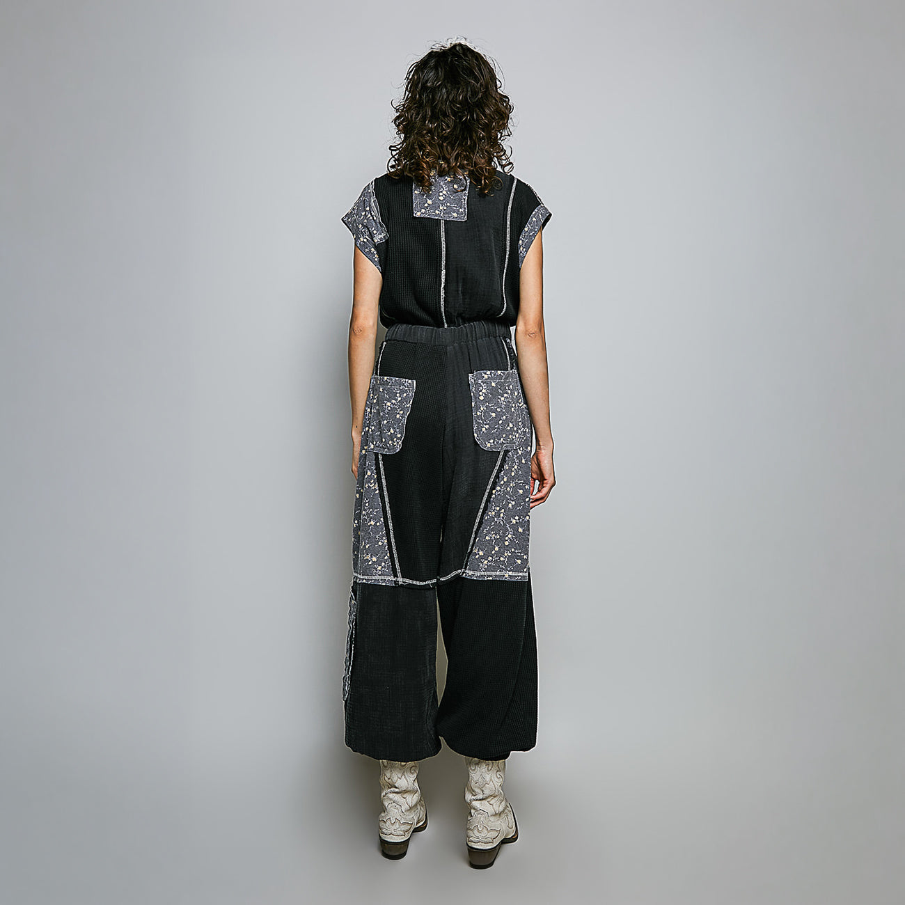 A model viewed from the back wearing a black wide-leg jumpsuit with silver patterned contrast panels and white lace-up boots, set against a plain light gray studio background.