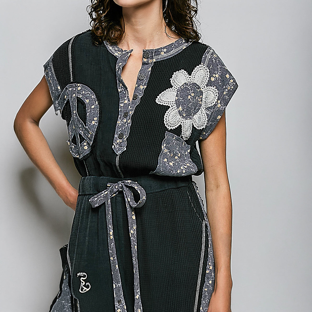 A woman wearing a dark green ribbed dress with gray patterned lace appliques, a peace sign on the chest, and a large flower motif, paired with a matching waist tie belt.