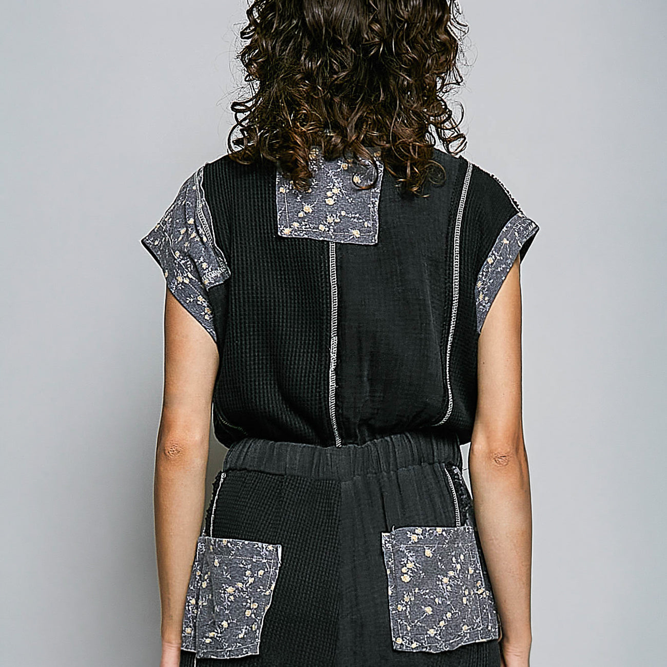 A model shown from the back wearing a black textured waffle-knit top and skirt set with gray floral patterned patchwork details on the collar, sleeve cuffs, and pockets, set against a plain gray studio background.