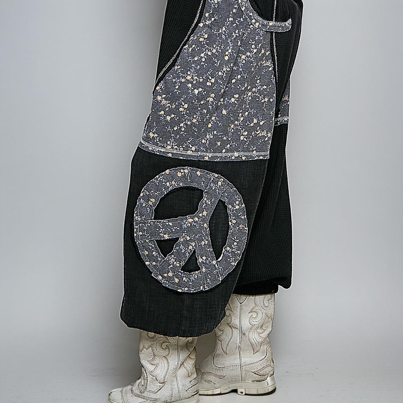 A close-up of a person wearing a black corduroy skirt or dress with a large gray peace sign patch and a gray floral fabric panel, paired with white western boots against a light gray studio background.
