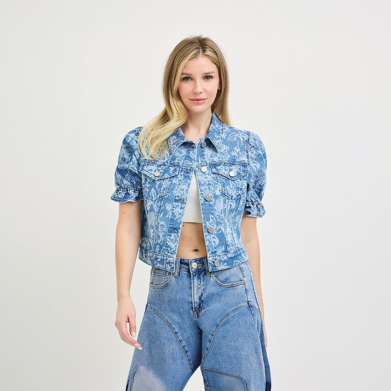 A blonde model wearing a cropped, short-sleeved blue denim jacket with a white abstract pattern, paired with high-waisted blue jeans, against a plain light gray studio background.