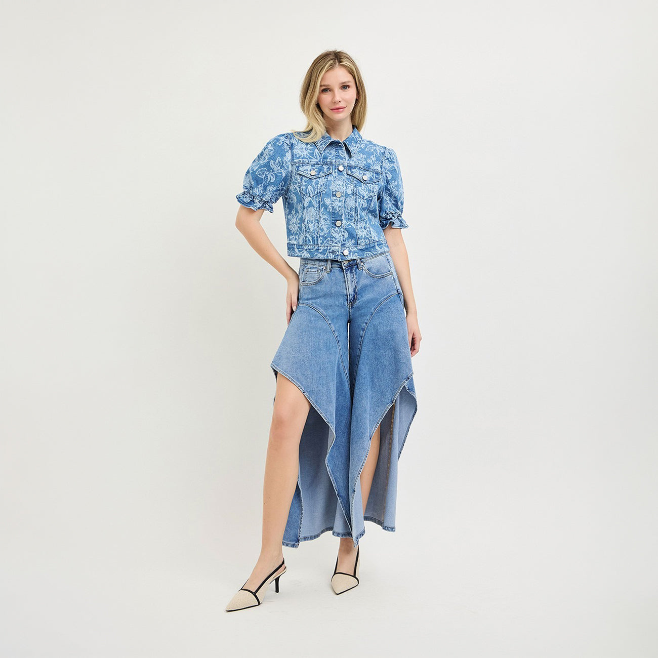 A woman modeling a blue floral short-sleeve button-up shirt paired with a high-low blue denim skirt and beige pointed-toe heels, set against a plain light gray studio background.