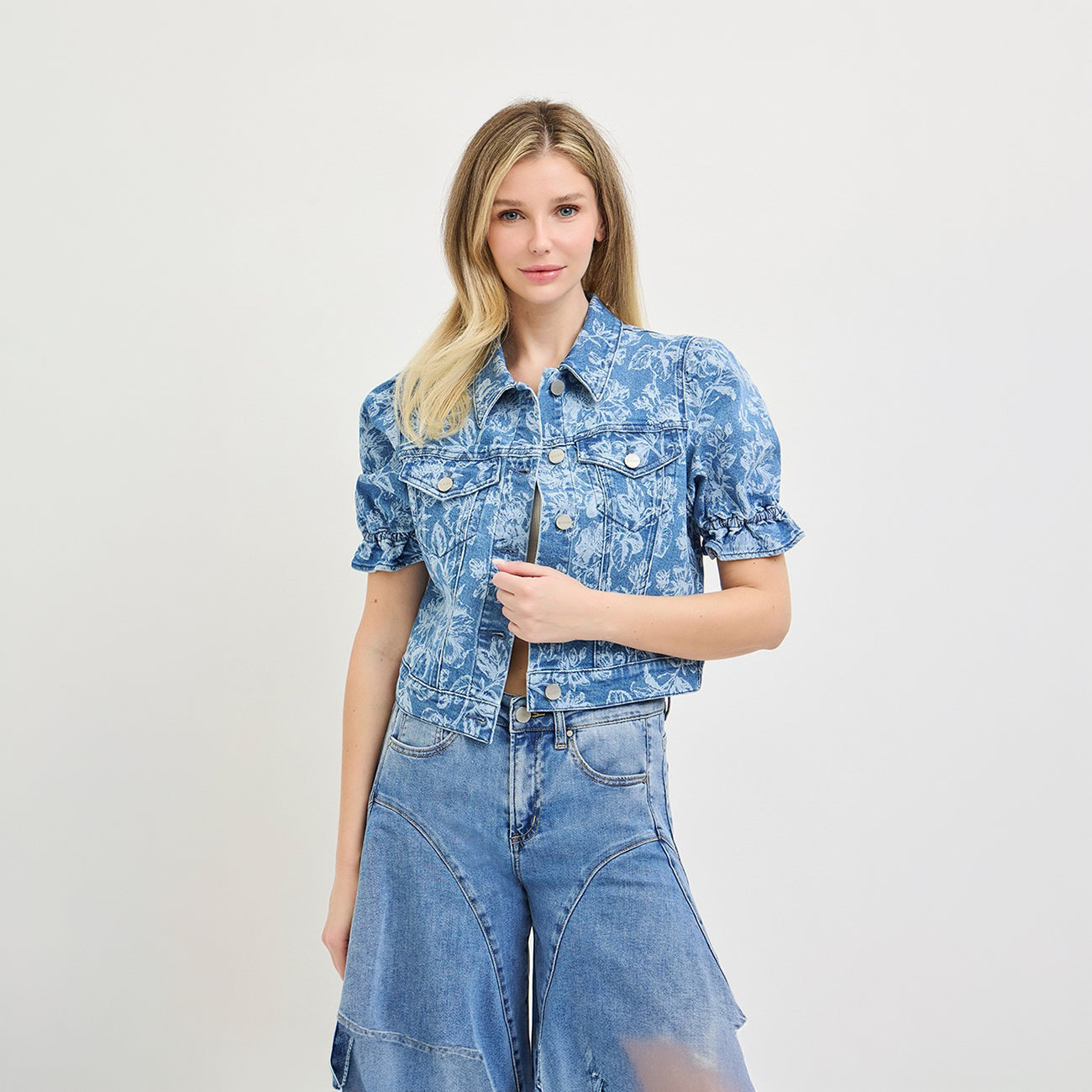 A blonde model wearing a short-sleeve blue and white floral print button-up shirt with ruffled sleeves, paired with high-waisted wide-leg denim jeans, against a plain light gray studio background.