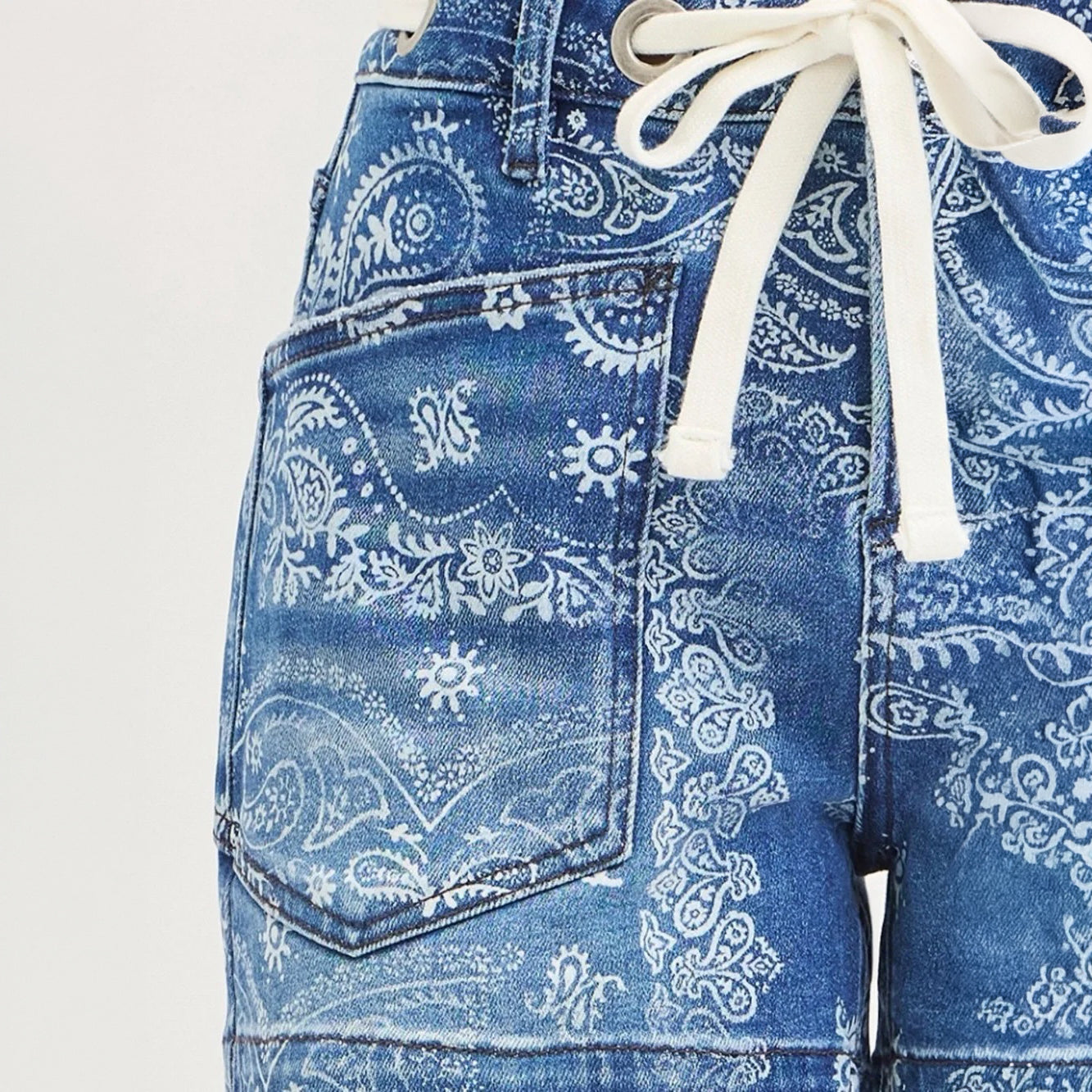 Close-up of blue denim shorts with a white paisley bandana print and a white drawstring waist, shown against a light neutral background.