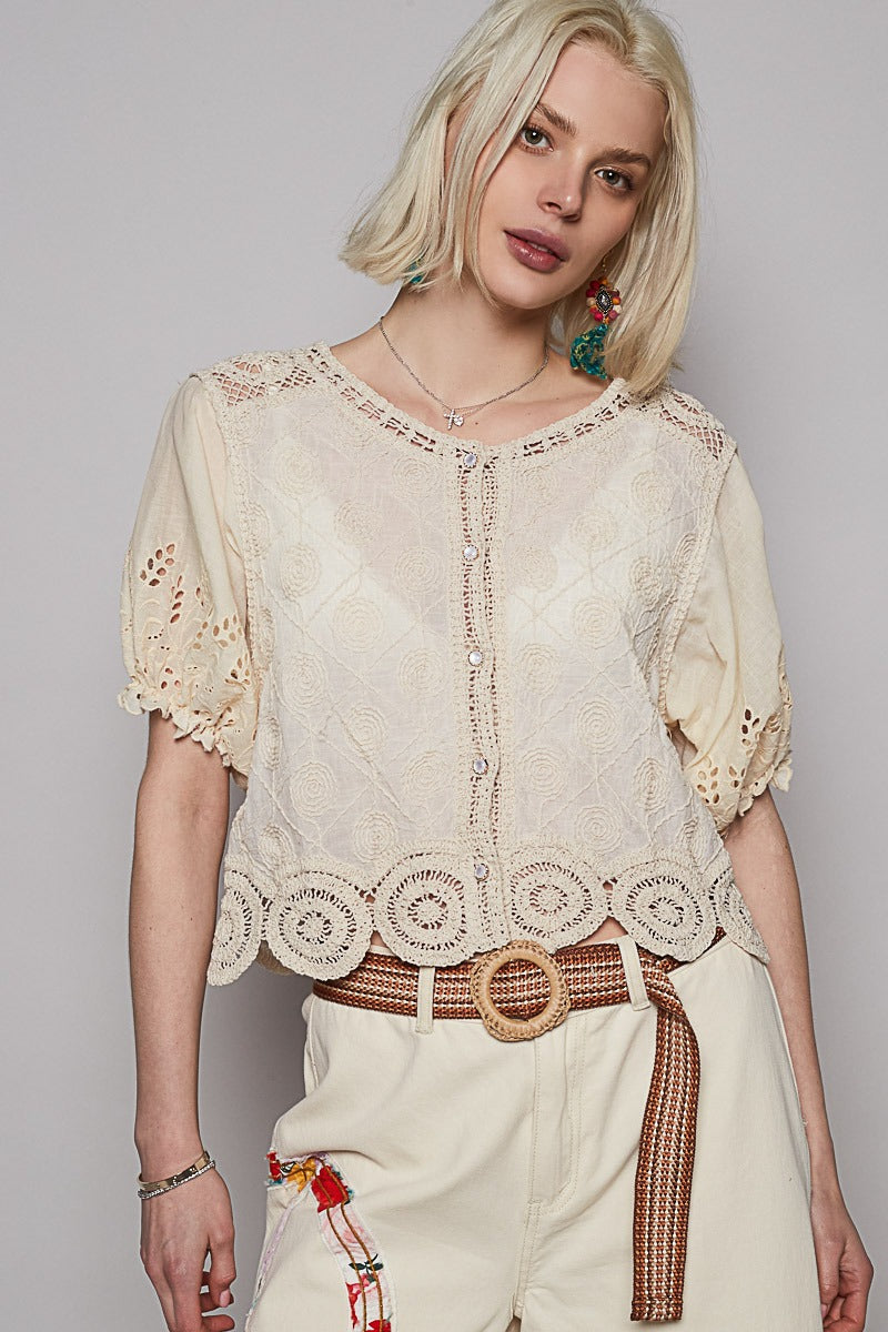 Woman wearing a beige lace top and white pants with a belt against a gray background