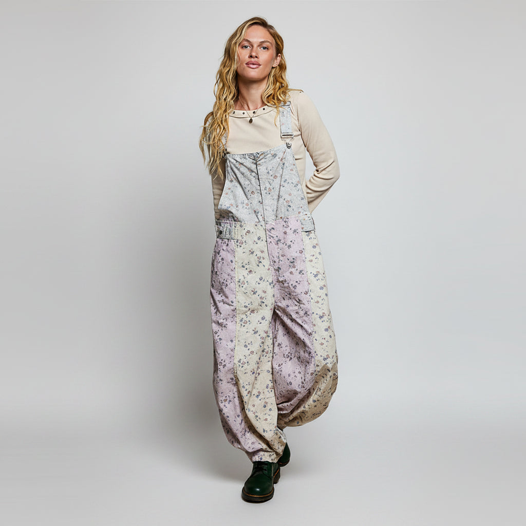 A woman modeling a pair of loose, balloon-style overall jumpsuits with a light lavender and beige floral patchwork print, worn over a cream long-sleeve top and dark green boots, set against a plain white background.