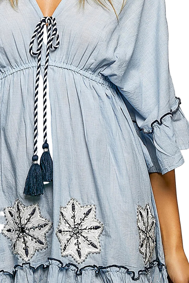 Close-up of POL Clothing light blue mini dress showing embroidered floral patches, rope tassel tie, and ruffle trim detail

