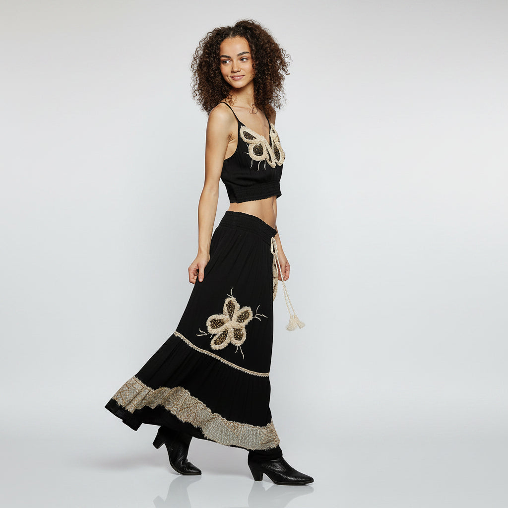 A woman modeling a black two-piece outfit consisting of a sleeveless crop top and a long skirt, both decorated with beige floral embroidery and beadwork, set against a plain white background.