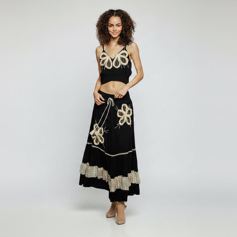A woman modeling a black two-piece dress set with intricate gold and white floral embroidery, featuring a sleeveless crop top and a long tiered skirt, set against a plain white background.