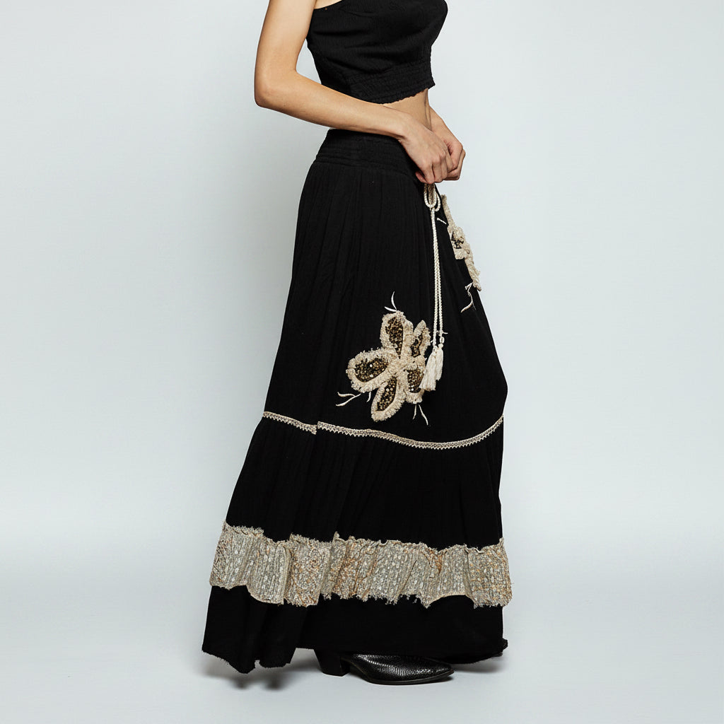 A black maxi skirt with a tiered ruffle hem, featuring a large butterfly embellishment and a decorative metallic embroidered band near the bottom, shown on a model against a white background.