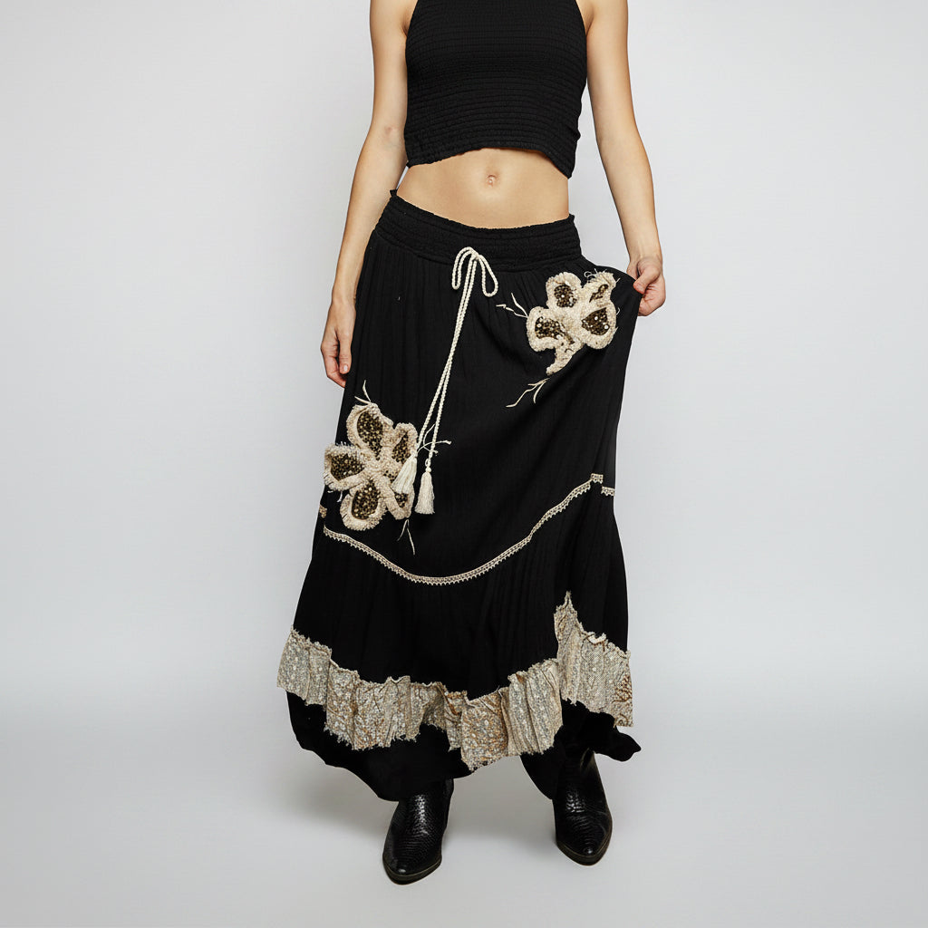 A black maxi skirt with intricate gold and beige floral embroidery, a white drawstring, and a ruffled gold lace hem, modeled against a white background.