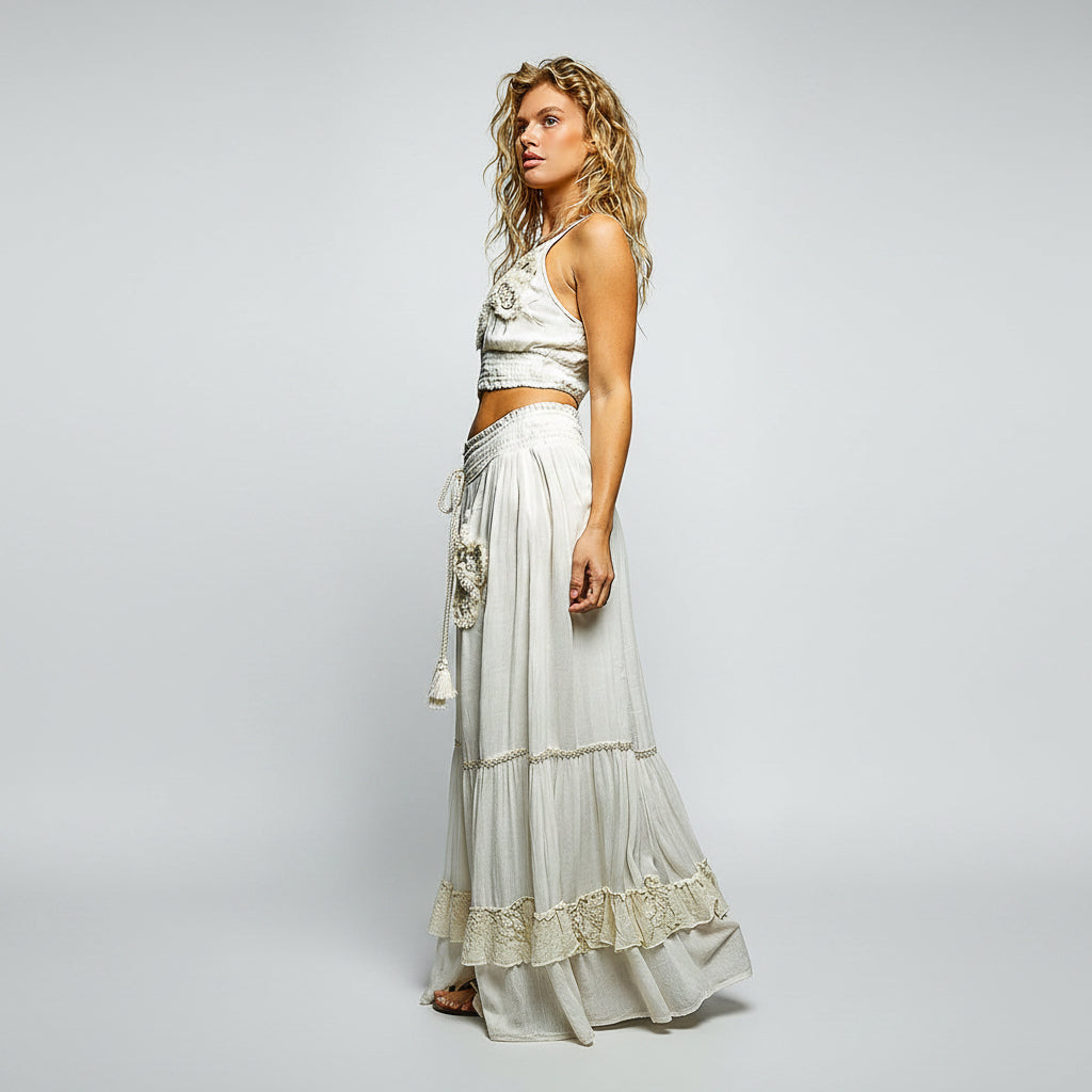 A woman modeling a bohemian-style two-piece outfit consisting of a white sleeveless crop top with floral embroidery and a matching tiered white maxi skirt with lace trim and a tassel tie belt, set against a plain white background.