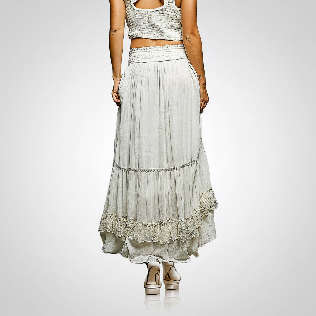 A woman modeling a long, tiered maxi skirt in a light sage green or gray-green chiffon fabric with lace trim, paired with a matching crop top, shown from the back against a plain white background.