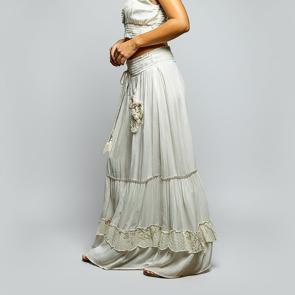 A woman wearing a light gray bohemian-style two-piece set consisting of a sleeveless crop top and a tiered maxi skirt with lace trim and a decorative hanging tassel belt, shown against a plain white background.