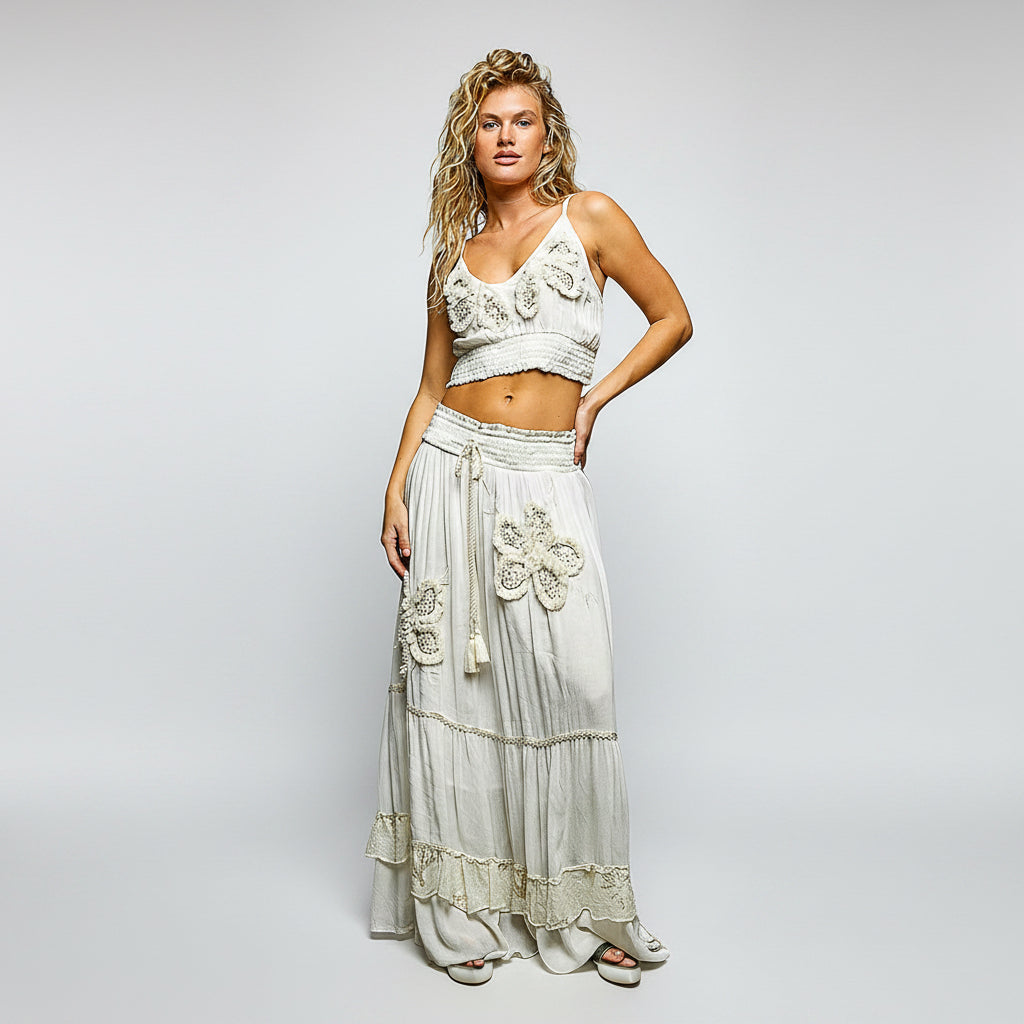A woman modeling a bohemian-style two-piece outfit featuring a white embroidered crop top and a matching long tiered maxi skirt with floral embroidery and tassels, set against a plain white background.
