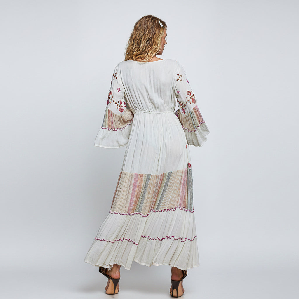 A woman viewed from the back wearing a long white boho maxi dress with colorful floral embroidery on the sleeves and a striped multicolor band near the hem, set against a plain white background.