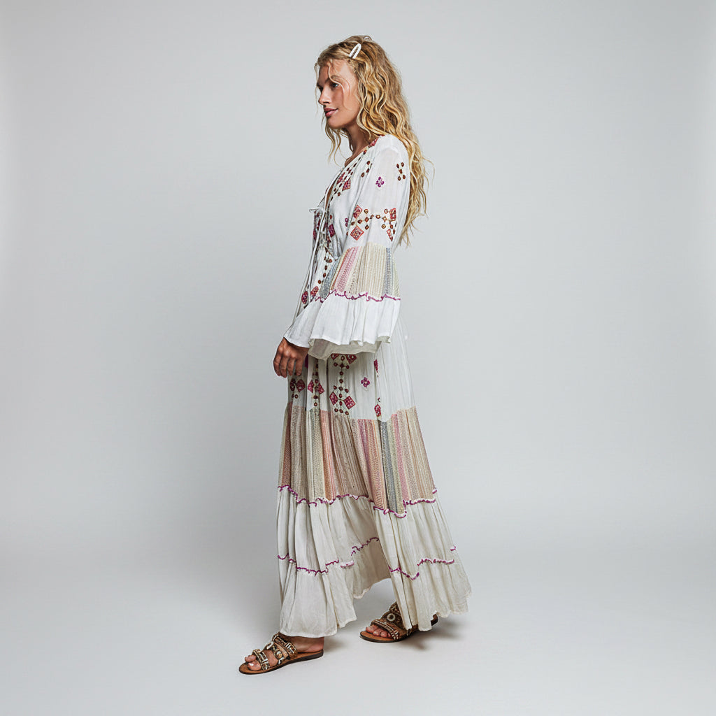 A woman modeling a long white bohemian maxi dress with colorful floral embroidery and tiered ruffles, shown from a side profile against a plain white background.