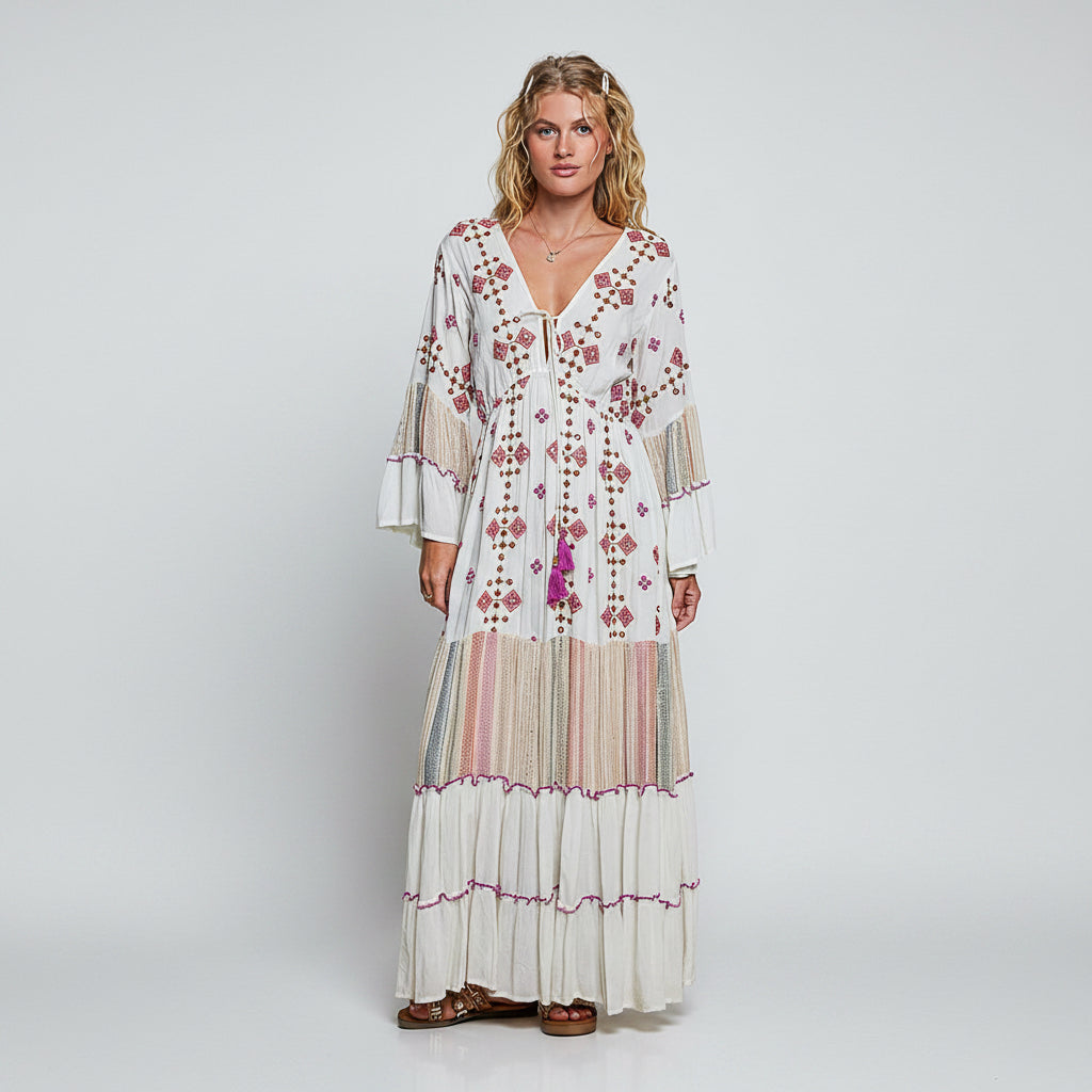 A woman modeling a long white bohemian maxi dress with pink floral embroidery, tiered ruffles, and striped fabric panels on a solid white background.