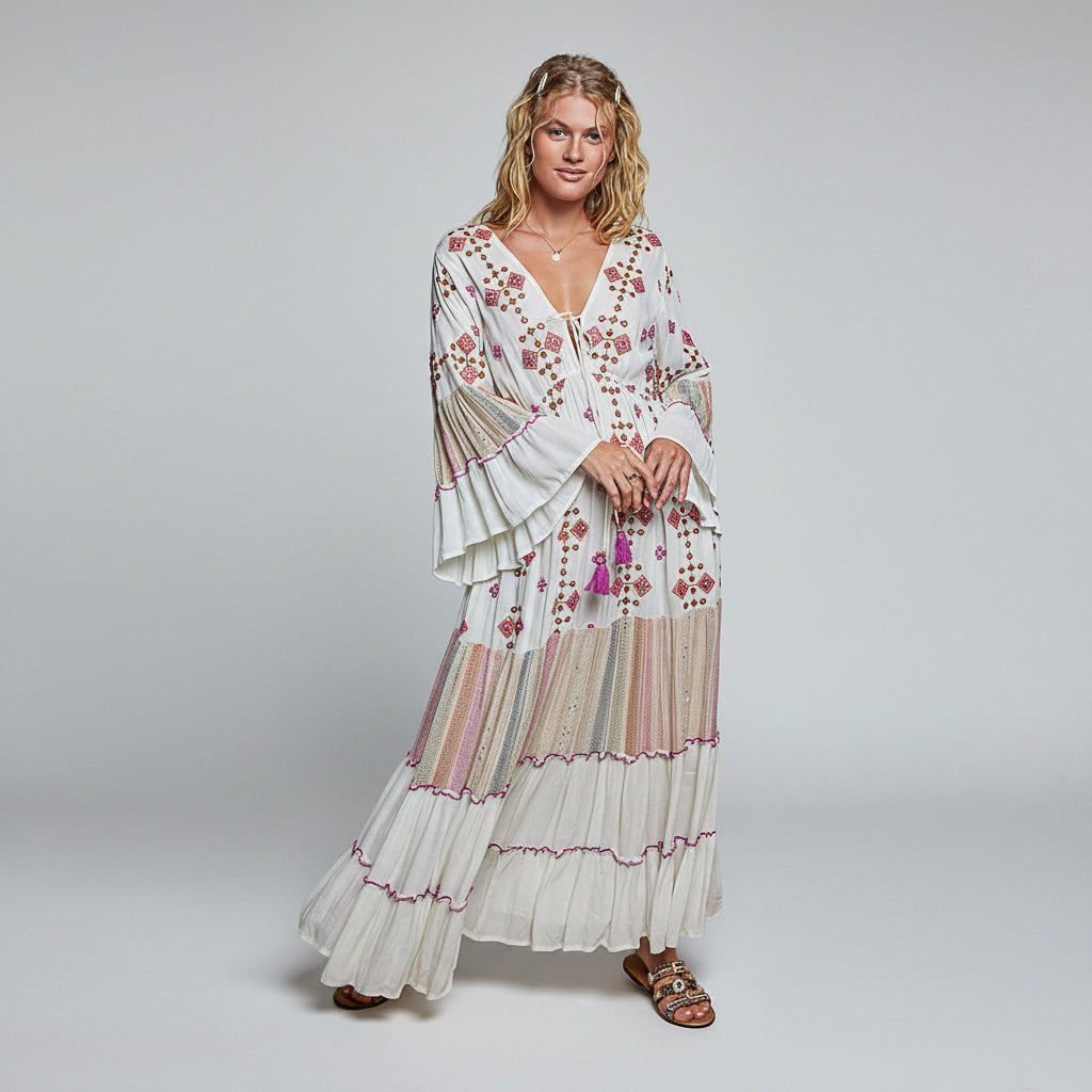 A woman modeling a long white bohemian maxi dress with pink and purple floral embroidery, tiered ruffle sleeves, and a tiered skirt, set against a plain white background.