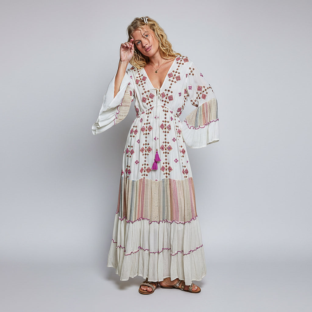 A woman modeling a bohemian-style maxi dress with a white base, pink and beige floral embroidery, and tiered ruffles, set against a plain white background.