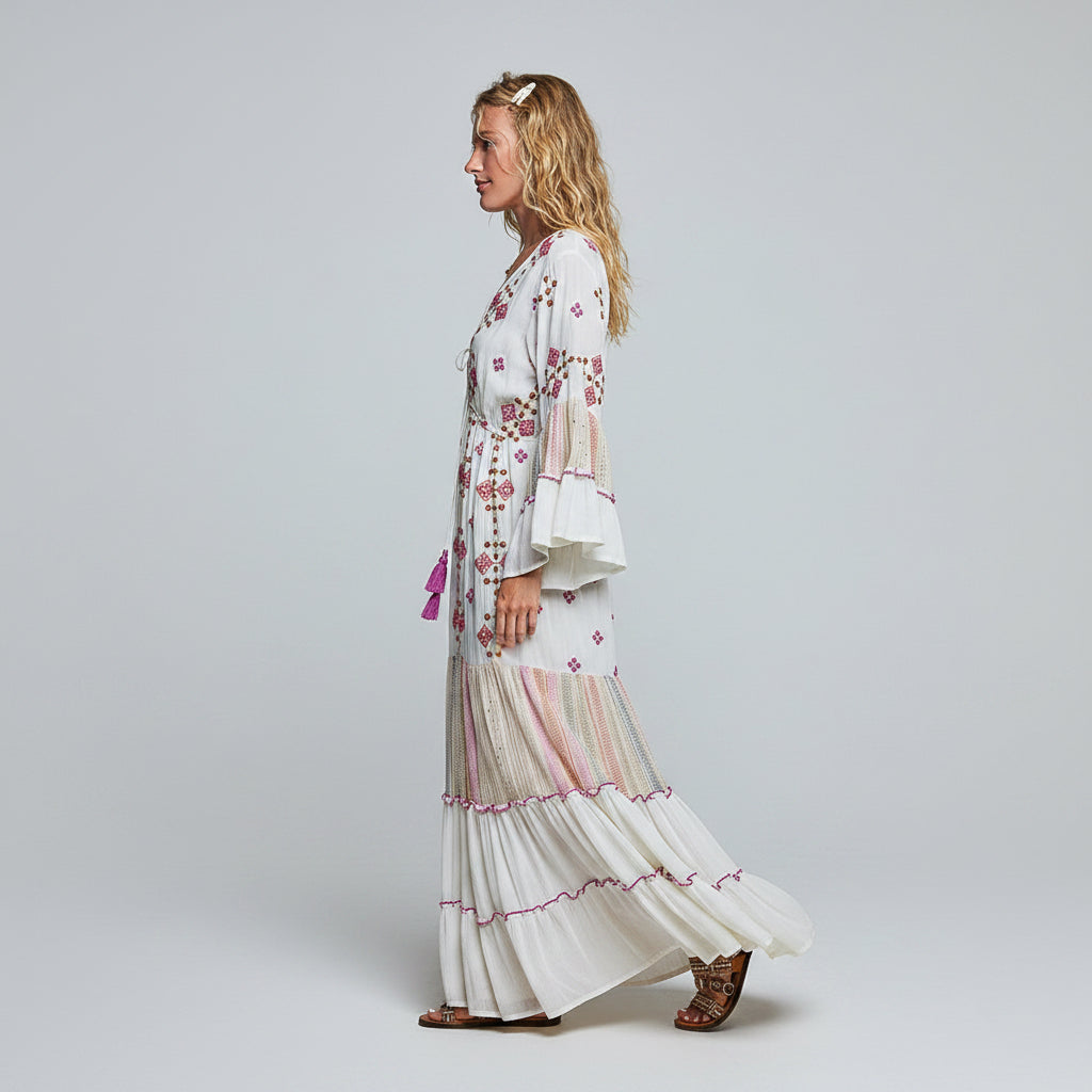 A woman modeling a long white bohemian maxi dress with pink and beige striped tiers, floral embroidery, and flared bell sleeves against a plain white background.