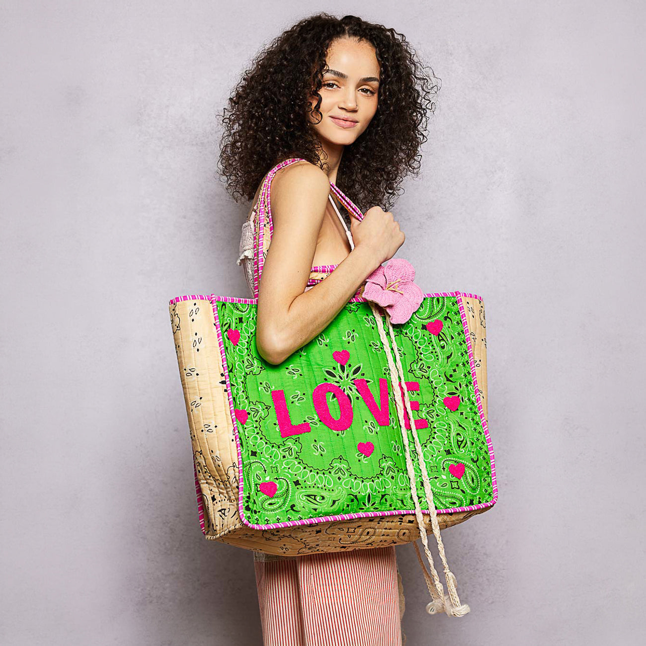 A woman with curly dark hair holds a large woven tote bag. The bag has a bright green front with pink 'LOVE' lettering, pink heart motifs, and pink trim, with a natural tan woven body and white rope handles.